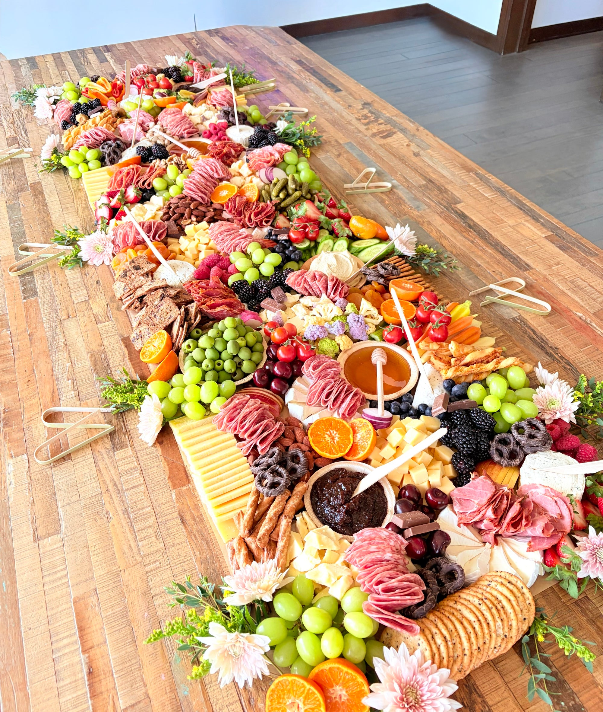 A large charcuterie board spread arranged diagonally on a wooden table, featuring sliced meats, cheeses, grapes, crackers, fruit, and small flowers.