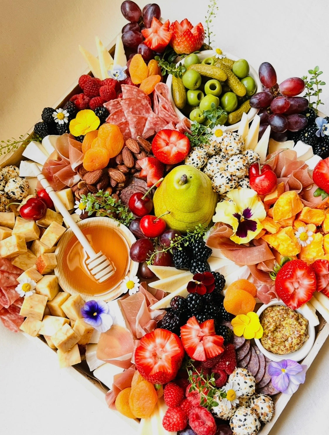 Assorted fruits, cheeses, and meats on a platter with flowers and honey dipper.