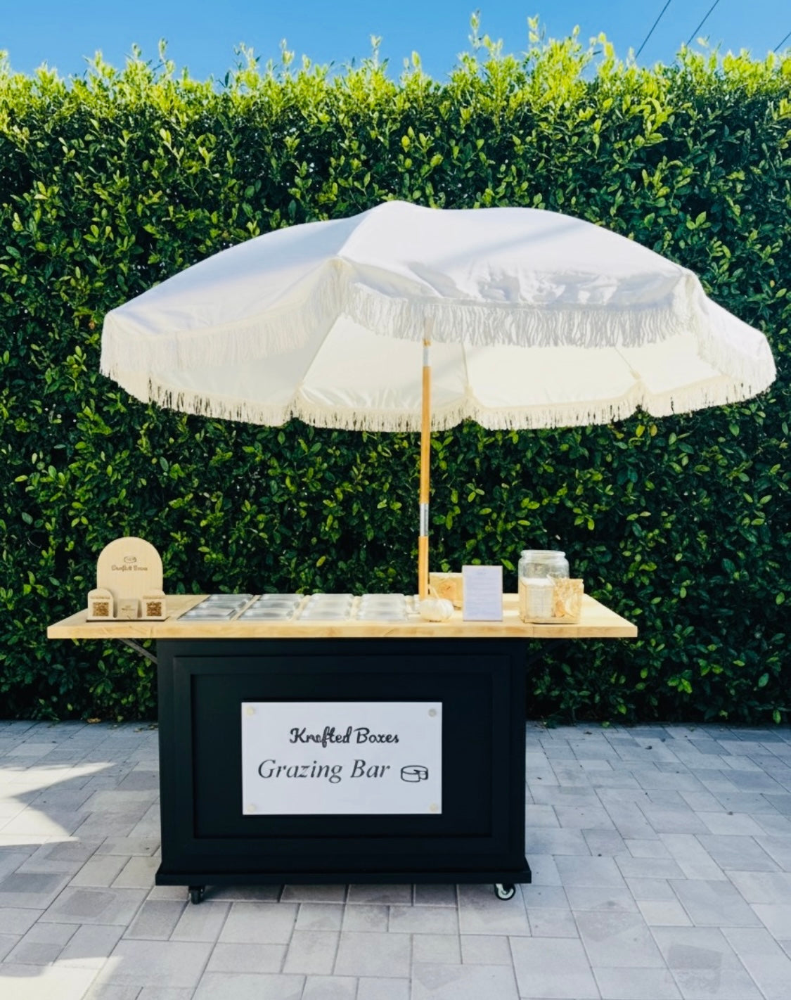 Grazing bar with white umbrella and snacks against a green hedge
