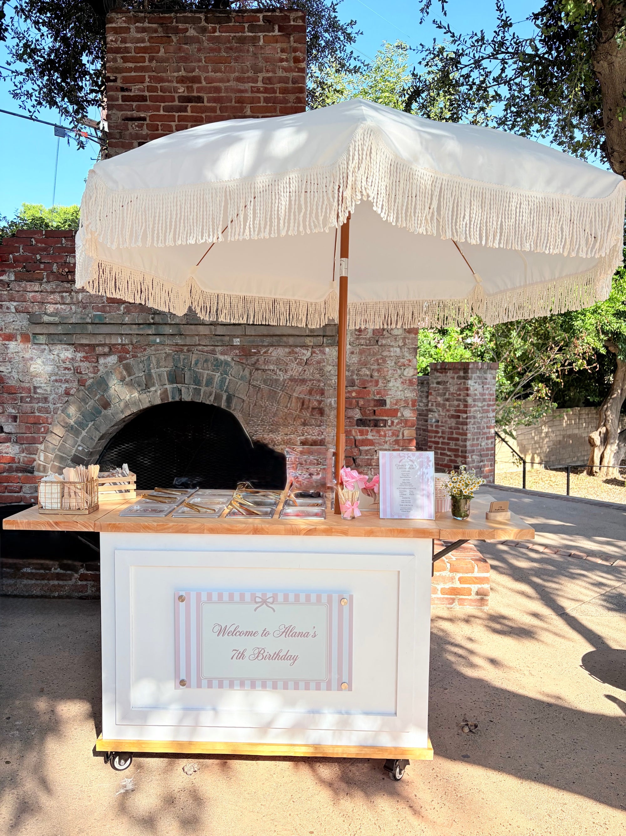A white rolling cart with a large cream fringe umbrella overhead, set outdoors in front of a brick oven. The cart has a wooden tabletop and a white front panel with a decorative sign that reads 'Welcome to Elena's 7th Birthday'.