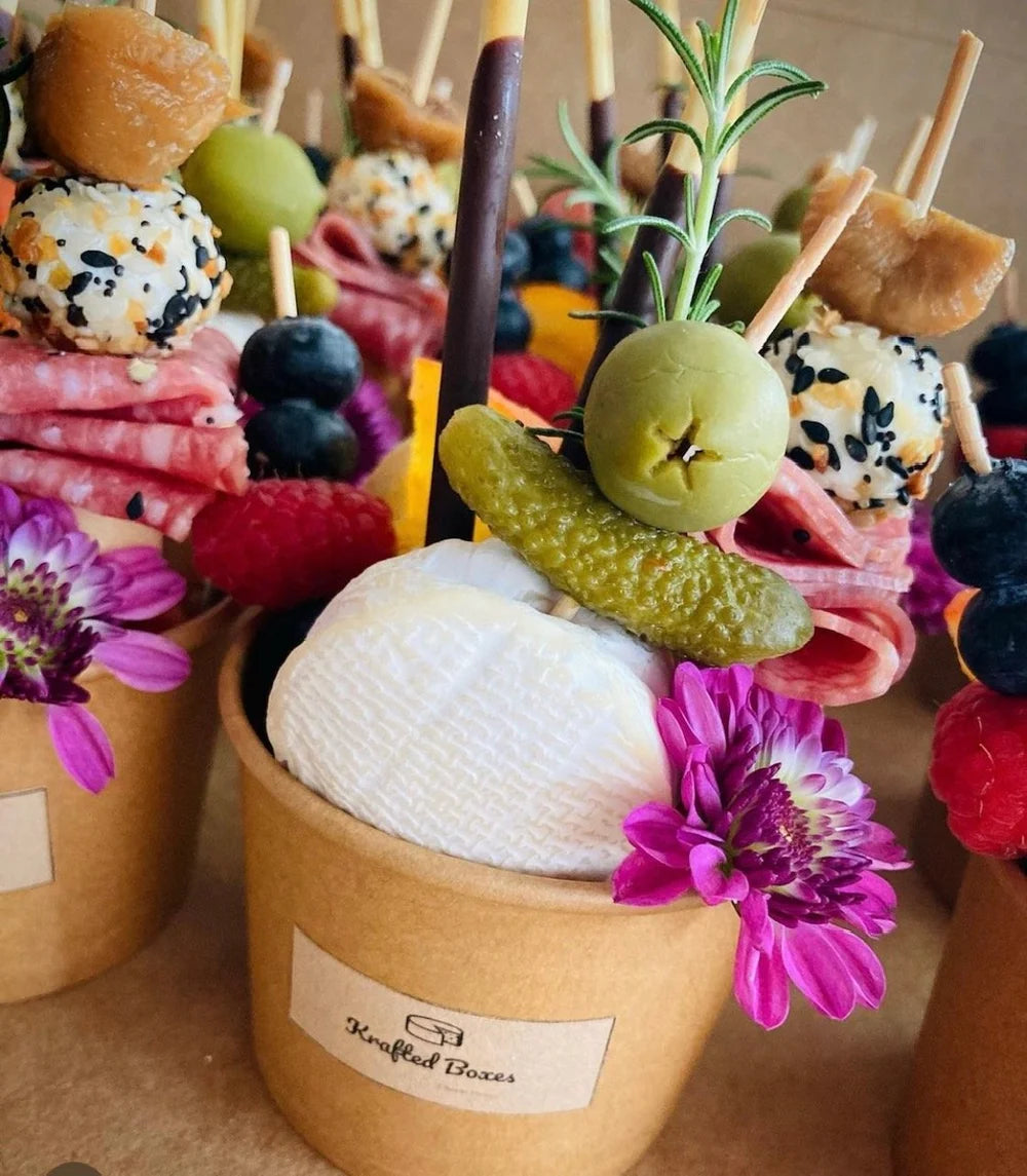 Decorative skewers with pickles, flowers, and other items in a brown container labeled 'Krafted Boxes'.