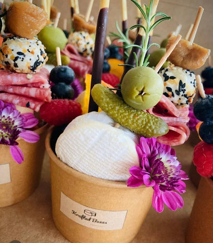 Decorative skewers with pickles, flowers, and other items in a brown container labeled 'Krafted Boxes'.