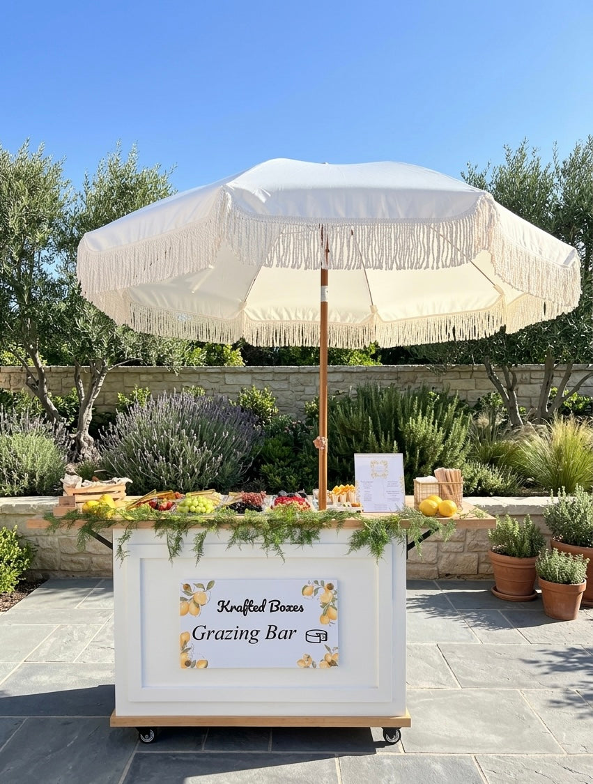 An outdoor grazing bar setup with a white fringed umbrella over a white rolling cart labeled 'Grassfed Beef Grazing Bar,' set on a stone patio with plants and trees in the background.